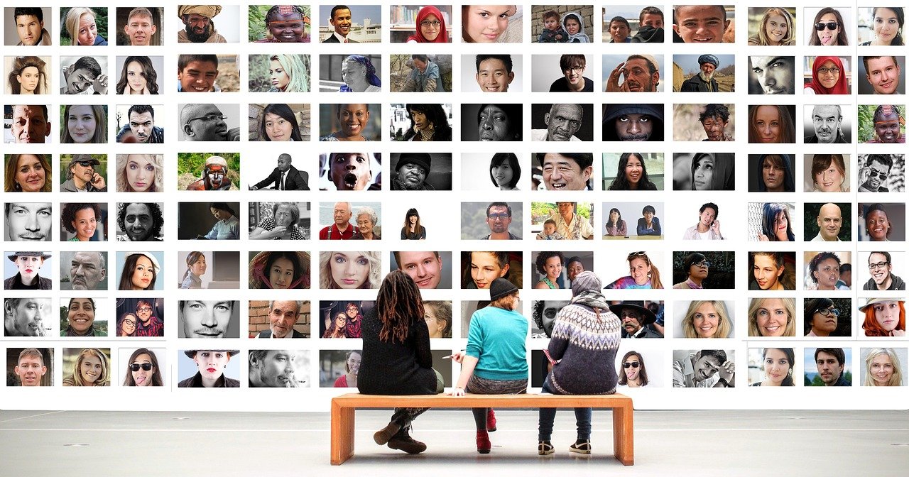 Three people sat on a bench in an art gallery. They are looking at a wall full of portraits